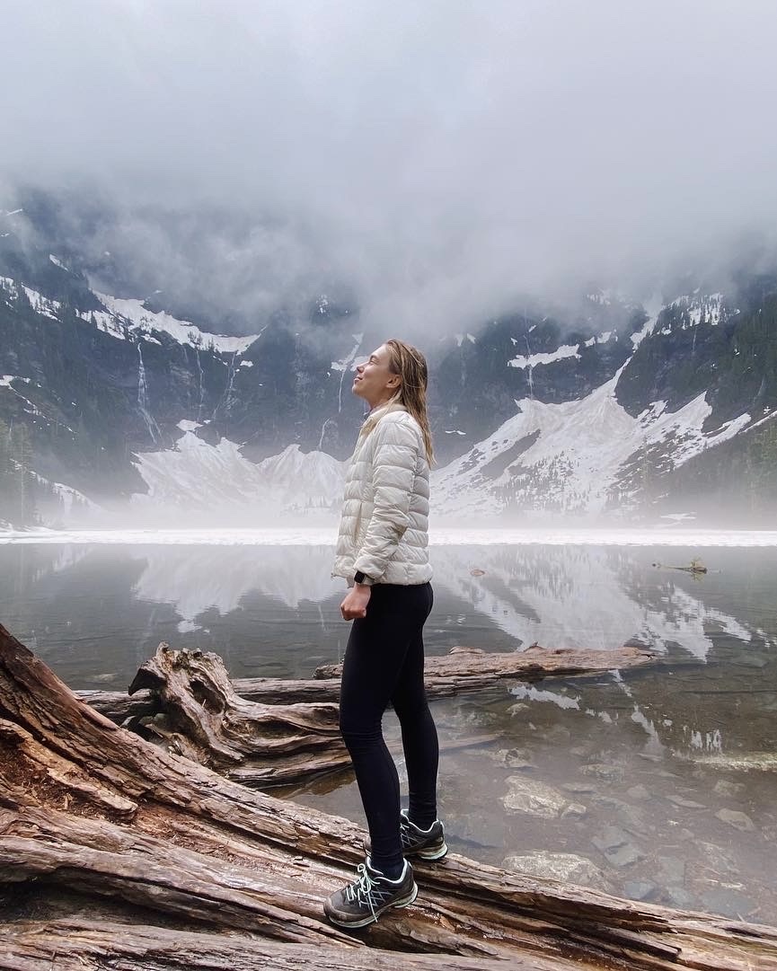 Girl standing on a piece of wood in front of the lake and mountains looking away.